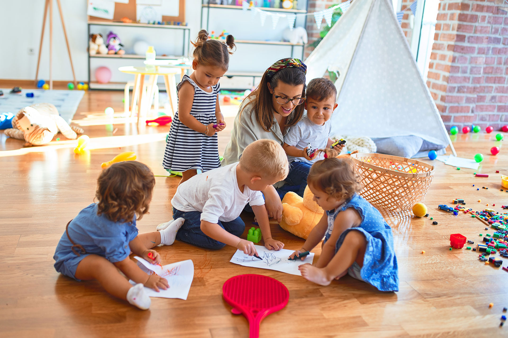 Teacher and group of toddlers sitting on the floor drawing using paper and pencil around lots of toys at an in-home daycare.