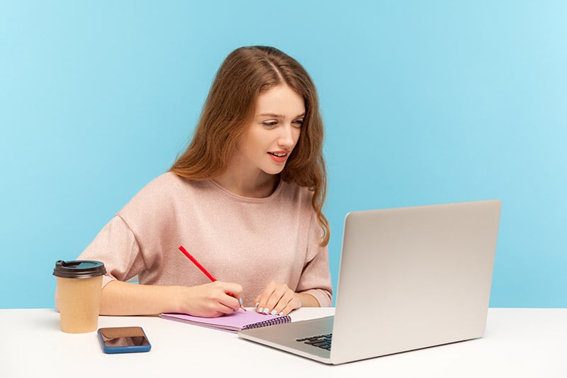 Woman taking notes as she looks at her laptop screen