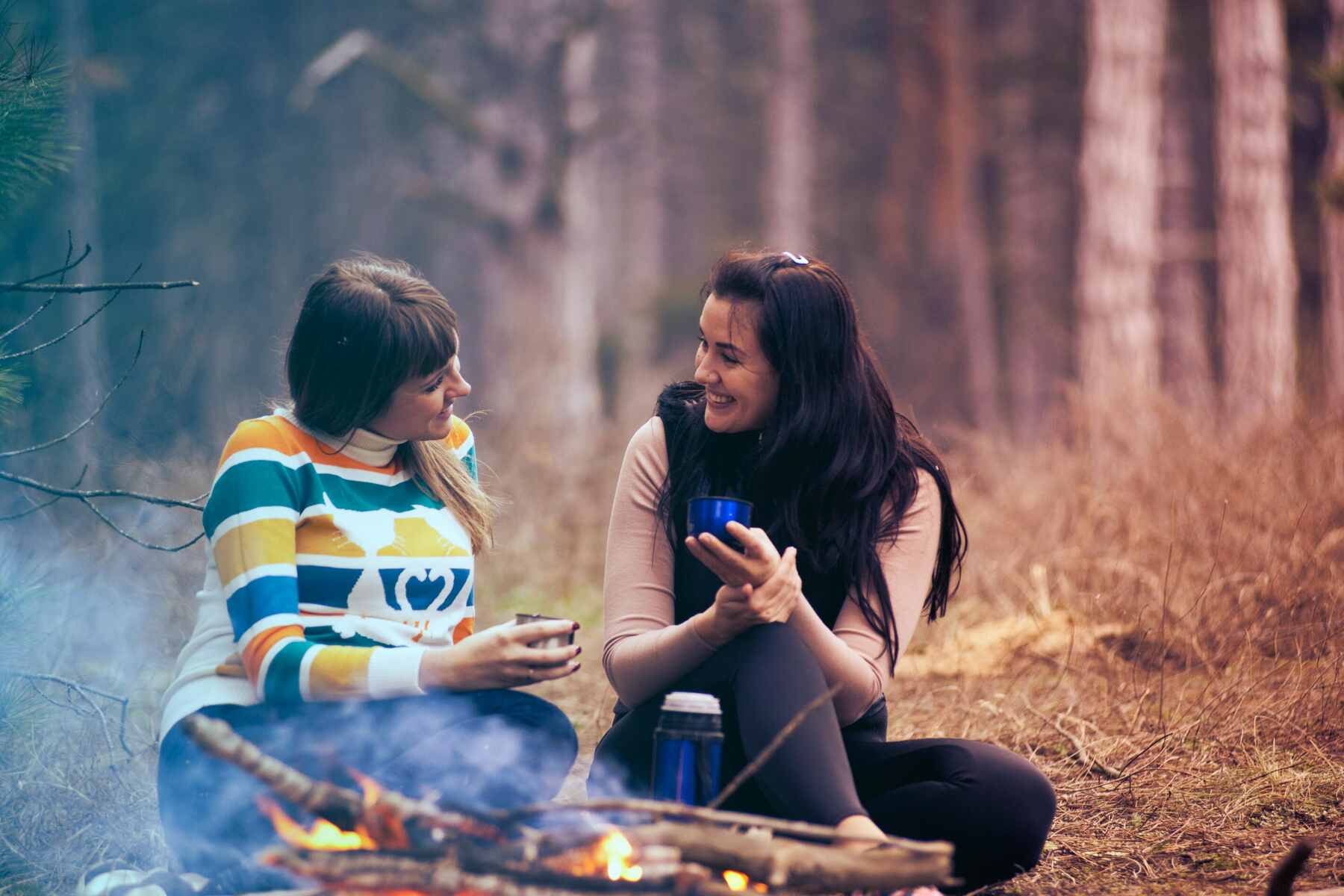 Two women holding thermos cups while sitting and talking to each other on a camp fire