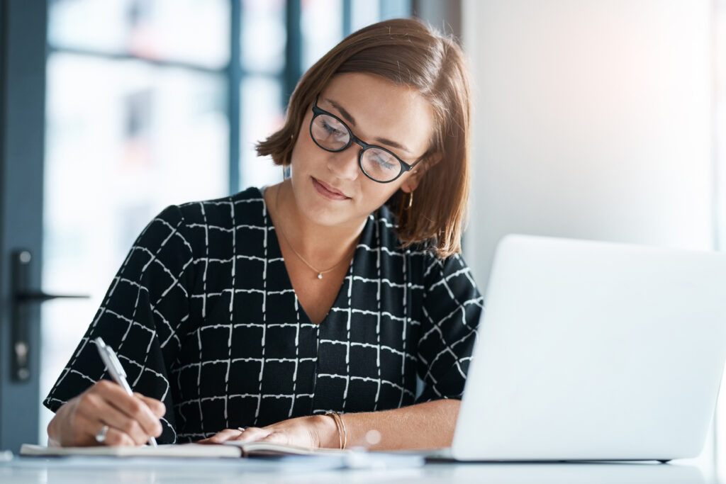 Professional woman writing in a notebook while working on a laptop in a modern office.