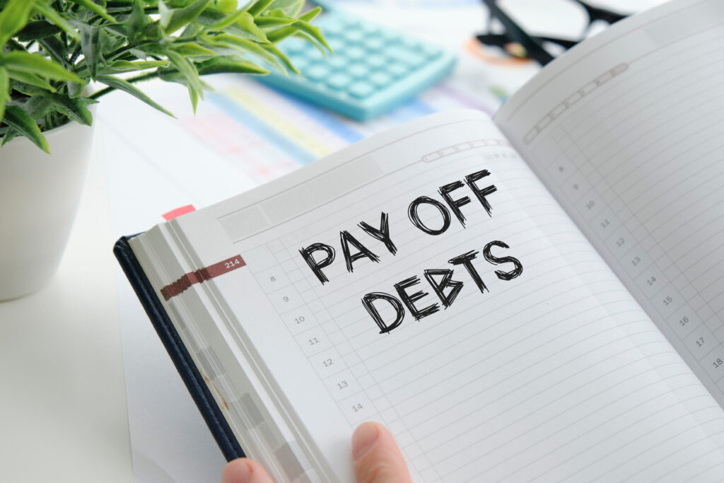 An open planner with the words "Pay Off Debts" written in bold, black font, surrounded by office supplies like a calculator, glasses, and a plant.