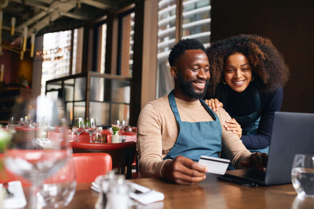 Pair of small business workers reviewing something online with a laptop and credit card inside a dining area.