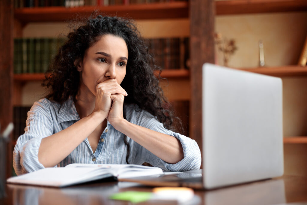 Woman sitting at a desk and looking thoughtfully at a laptop, with an open notebook in front of her.